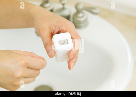 Foto orizzontale di mani femminili tirando il filo interdentale al di fuori del contenitore in bagno con lavandino e bancone in background Foto Stock