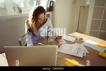 Piuttosto giovane femmina note di scrittura sorridente. Bella femmina caucasica con computer portatile lavora da casa in ufficio. Foto Stock