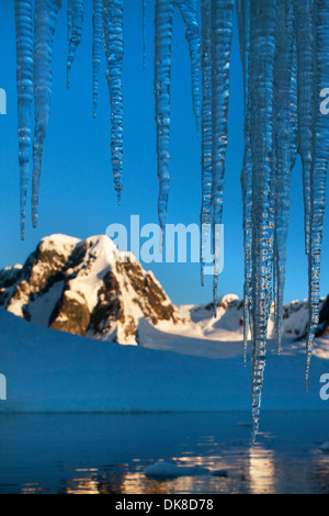 L'Antartide, Petermann Island, impostando il sole illumina le montagne sopra Lemaire Channel dietro ghiaccioli appesi da iceberg Foto Stock