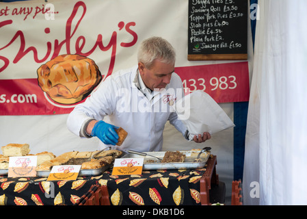 Uomo che indossa mantello bianco e indossando guanti blu il prelievo di torta a base di carne di servire il cliente sul mercato Derbyshire stallo Inghilterra Foto Stock