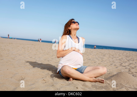 Donna incinta seduta sulla spiaggia Foto Stock