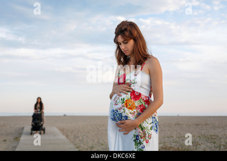 Donna incinta in piedi sul Boardwalk, donna spingendo passeggino in background Foto Stock