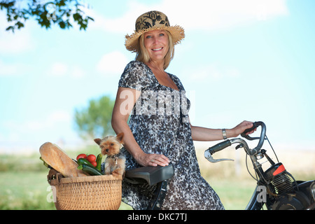 Donna matura sulla bicicletta elettrica con il cane e le verdure nel cestello Foto Stock