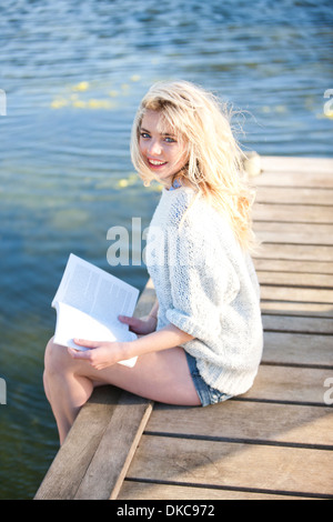 Giovane donna seduta sul molo azienda prenota Foto Stock