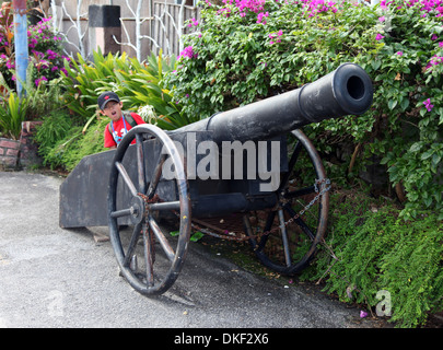 Si tratta di una foto di un vecchio cannone della guerra che stare vicino a una siepe in un cortile di un museo in Vietnam. Un ragazzo si finge di utilizzarlo Foto Stock