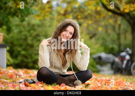 Sorridente giovane donna seduta in foglie di autunno Foto Stock