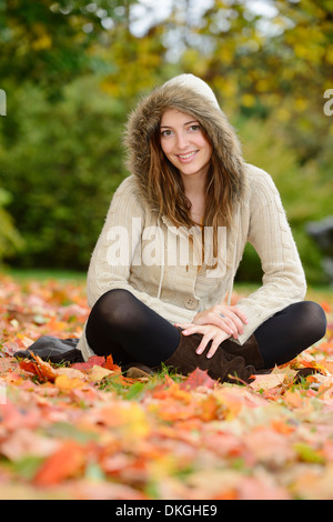 Sorridente giovane donna seduta in foglie di autunno Foto Stock