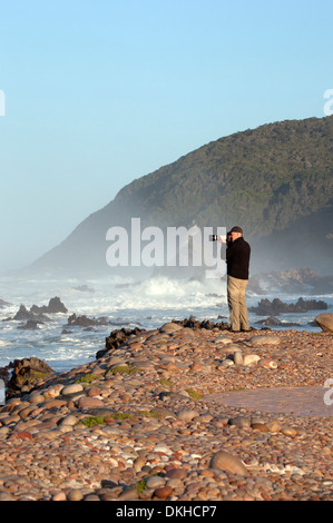 Soleggiato turista maschio sulla ghiaia a fotografare l'oceano indiano a tsitsikamma national park in Sud Africa Foto Stock