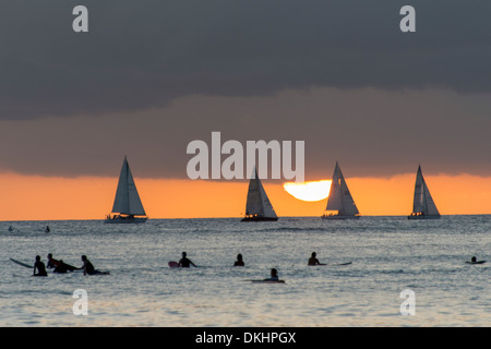 Surfisti sulla spiaggia con barche a vela in background al tramonto, Waikiki, Honolulu Oahu, Hawaii, STATI UNITI D'AMERICA Foto Stock