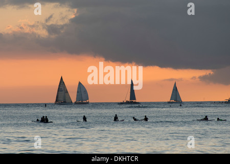 Surfisti sulla spiaggia con barche a vela in background al tramonto, Waikiki, Honolulu Oahu, Hawaii, STATI UNITI D'AMERICA Foto Stock