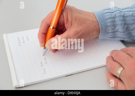 Parte superiore verso il basso di una mano destra senior donna di mano che tiene una penna a scrivere una lettera su un blocco per appunti di carta su un tavolo. Inghilterra, Regno Unito Foto Stock