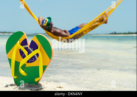 L uomo si rilassa in amaca sul mare lasciando la sua bandiera Brasile flip flop nella sabbia Foto Stock