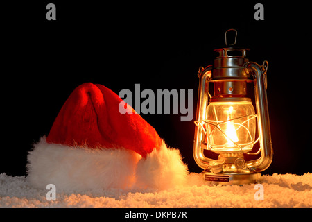 Una Santa Claus o Babbo Natale hat sulla neve illuminata dalla luce di un olio riempito lanterna, sfondo nero. Foto Stock