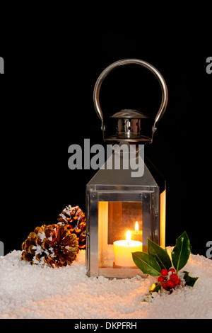 Un Natale lanterna con una candela brucia in neve con holly e pigne, sfondo nero. Foto Stock