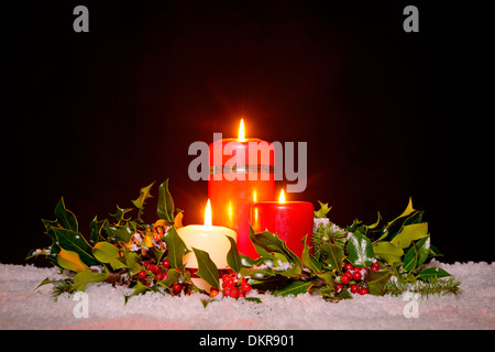 Natale ancora la vita di tre candele sulla neve circondato da un verde e fresco holly, edera e ghirlanda di abete rosso Foto Stock