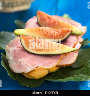 La figura freschi fette di prosciutto affumicato panino sulle foglie di fico su legno blu (messa a fuoco selettiva, Focus sulla prima fetta di figura) Foto Stock