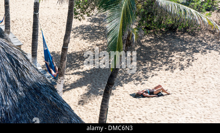 Visualizza in basso uomo sdraiati in amaca & donna ensoleillement se stessa al di fuori hotel sul fronte spiaggia sul palmo rivestito idilliaco San Agustinillo beach Foto Stock