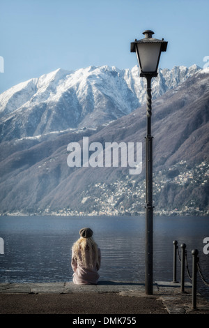 Una ragazza in un cappotto rosa è seduto di fronte ad un lago, sullo sfondo delle montagne coperte di neve Foto Stock