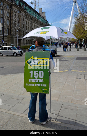 Subway sandwich shop un'attività di vendita al dettaglio pubblicizzata su un uomo che trasporta panini in piedi sul marciapiede con il London Eye oltre l'Inghilterra Regno Unito Foto Stock
