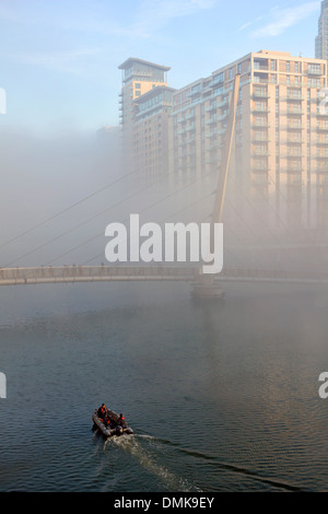 La nebbia la cancellazione da Canary Wharf e vie navigabili con piccola barca passando sotto la gente camminare su passerella rottura sole attraverso Foto Stock