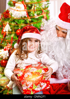 Poco felice ragazza ricevere il dono di Babbo Natale, seduti vicino al bellissimo albero di Natale decorato, godendo la vigilia di Natale a casa Foto Stock