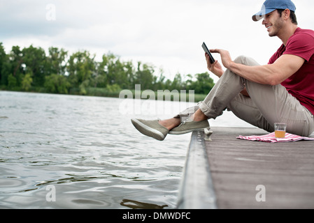 Un uomo seduto su di un molo da un lago utilizzando una tavoletta digitale. Foto Stock