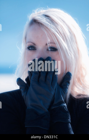 Ritratto di una giovane e bella donna con capelli lunghi biondi che tiene le mani con guanti neri nella parte anteriore della bocca Foto Stock