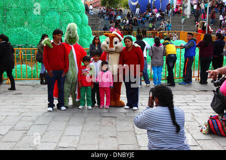 La Paz in Bolivia. Il 21 dicembre 2013. Una famiglia locale pone per foto istantanee con il Grinch e Alvin Scoiattolo striado in Plaza San Francisco. In fondo è ecologico di un albero di Natale fatto di plastica riciclata bottiglie di bevande. La struttura ad albero è superiore a 15m di altezza e contiene circa 50.000 bottiglie. Credito: James Brunker / Alamy Live News Foto Stock