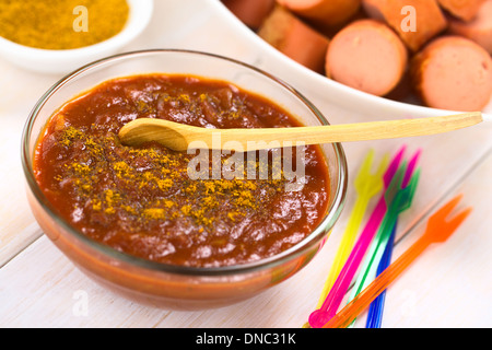 Curry ketchup salsa fatta immergere le salsicce fritte servite in recipiente di vetro con il cucchiaio di legno, plastica colorata parte forche sul lato Foto Stock