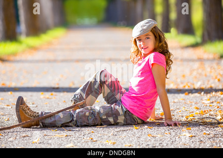 Autunno kid ragazza con Pantalone mimetico seduto appoggiato in caduta di alberi via con escursionista stick di diramazione Foto Stock