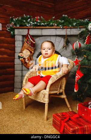 Piccolo Ragazzo seduto nella sedia di vimini vicino albero di Natale sullo sfondo di caminetti Foto Stock