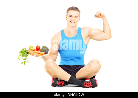 Giovane atleta maschio su un pavimento afferrando un piatto pieno di verdure fresche e mostrando il suo muscolo Foto Stock