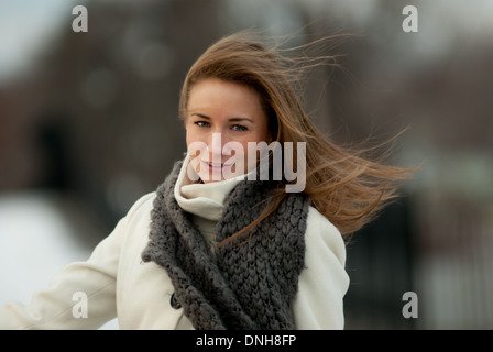 Una giovane e bella donna in un manto bianco e grigio sciarpa sorrisi su un ventoso giorno d'inverno. Foto Stock