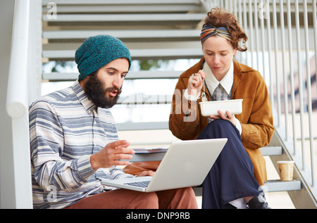 Hipster giovane utilizzando il computer e mangiare il pranzo seduti in scale al campus universitario Foto Stock