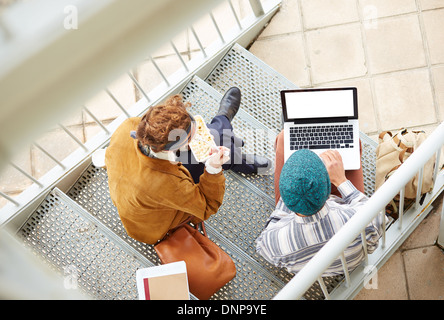 Hipster giovane utilizzando il computer e mangiare il pranzo seduti in scale al campus universitario Foto Stock