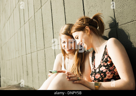 Giovani donne seduta e appoggiata contro la parete, guardando a smart phone insieme Foto Stock