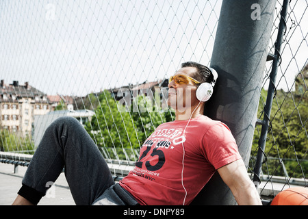 Uomo maturo seduti all'aperto sul campo di pallacanestro che indossa le cuffie e ascolto di musica, Germania Foto Stock