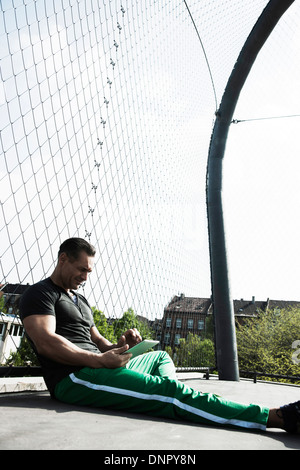 Uomo maturo seduto su outdoor pallacanestro, guardando il computer tablet, Germania Foto Stock