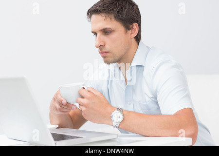 Uomo serio con tazzina utilizzando portatile a casa Foto Stock