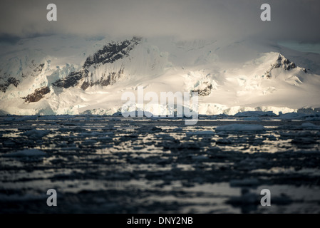 Paradise Harbour Antartide con iceberg e montagne al tramonto | PARADISE HARBOUR, Antartide — Gli iceberg e il ghiaccio glaciale riempiono lo sfondo di Paradise Harbour sotto montagne innevate che catturano l'ultima luce arancione della giornata sotto un cielo nuvoloso. La baia si trova sulla costa Danco della Terra di Graham, protetta dietro le isole Lemaire e Bryde, ed è uno dei pochi siti della penisola antartica dove le navi sbarcano i passeggeri direttamente sul continente. Foto Stock