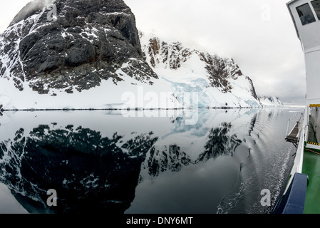 Riflessi del Canale Lemaire Punto Glandaz Antartide | CANALE LEMAIRE, Antartide — La prua di una nave e una passerella si inseriscono nel quadro mentre scogliere ripide e coperte di neve al Punto Glandaz si riflettono nelle acque specchianti di questo stretto passaggio antartico. Conosciuto come “Kodak Gap” per il suo paesaggio drammatico e fotogenico, la geografia ristretta del canale lo protegge dal vento che solitamente agita l’Oceano Australe circostante. Foto Stock