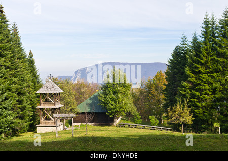 Punto di vista su un paesaggio del monte Bobija, prato davanti di una vecchia chiesa di legno circondata da alti alberi di abeti con picchi rocciosi Foto Stock