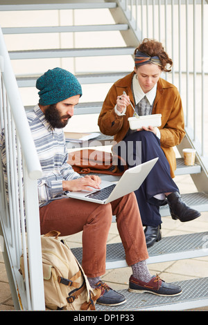 Hipster giovane utilizzando il computer e mangiare il pranzo seduti in scale al campus universitario Foto Stock