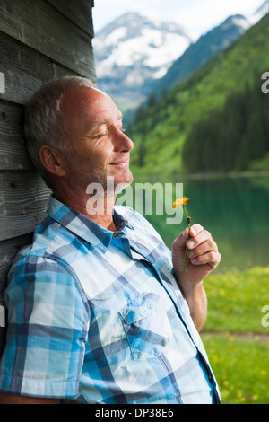 Ritratto di uomo maturo con gli occhi chiusi, tenendo fiore, in piedi accanto alla costruzione presso il lago Vilsalpsee, Valle di Tannheim, Austria Foto Stock