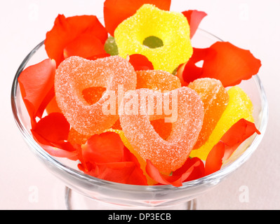Caramelle colorate con due cuori rossi in recipiente di vetro e petali di rose, close up Foto Stock