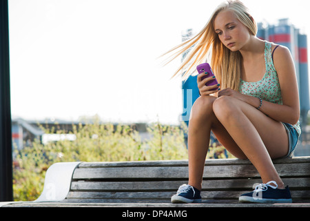 Ragazza seduta sul banco di prova all'aperto, guardando il telefono cellulare, Germania Foto Stock