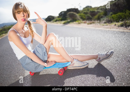 Funky bionda seduta sul suo skateboard rendendo rock e il gesto a mano Foto Stock