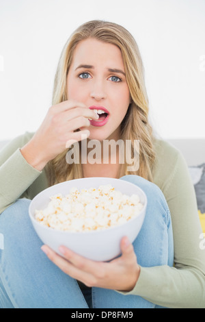 Sorpreso bella bionda mangiare popcorn seduto sul divano accogliente Foto Stock
