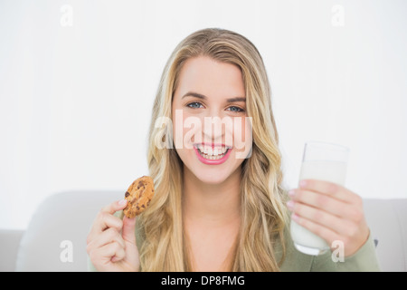 Sorridente biondo carino mangiare cookie con latte seduto sul divano accogliente Foto Stock