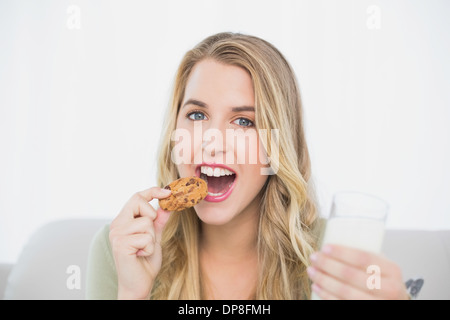 Allegro carino biondo mangiare cookie con latte seduto sul divano accogliente Foto Stock